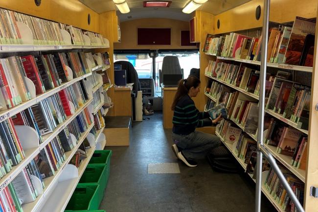 Inside of the Bookmobile with shelves of books and dvds and a patron holding and looking at books.